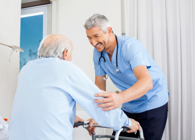 Smiling male caretaker helping senior man to use walking frame in bedroom at nursing home